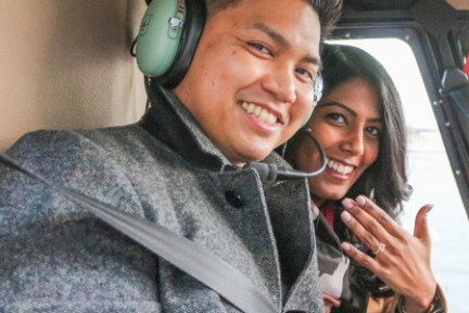 Smiling couple in helicopter, woman showing engagement ring.