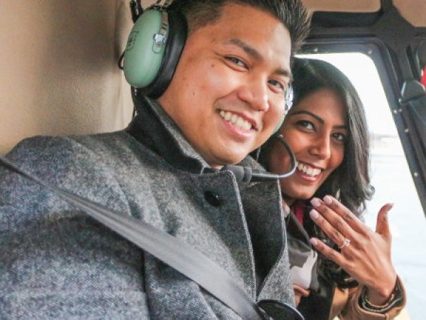 Smiling couple in helicopter, woman showing engagement ring.