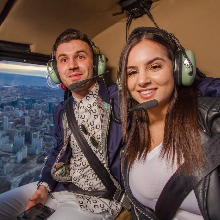 Couple wearing headsets in a helicopter above a cityscape.