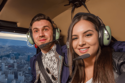 Two people smiling inside a helicopter with city view in the background.