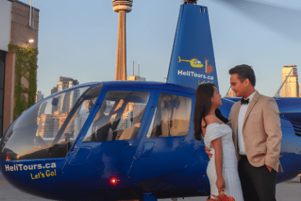 Couple in formal attire standing beside a blue helicopter with cityscape and tower in the background.
