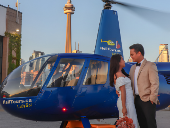 Couple in formal attire standing beside a blue helicopter with cityscape and tower in the background.